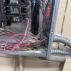 Electrical panel with pink and gray cables, breaker switches, and conduit connections mounted on exterior wall