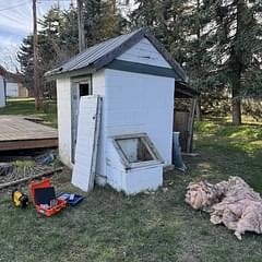 White and blue small shed or tiny house with dark roof in grassy yard surrounded by trees and landscaping rocks