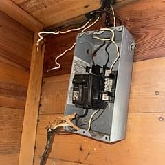 Open electrical panel mounted on wooden wall showing interior circuit breakers and wiring