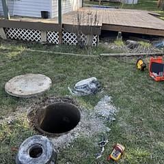 Open manhole in grassy yard with concrete covers and tools scattered nearby, white lattice fence in background