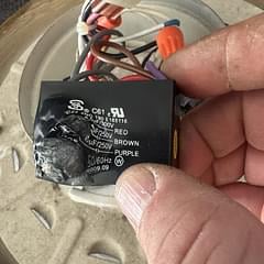 Hand holding a black capacitor with red and white wire terminals against a blurred background