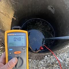 Hand holding digital multimeter showing 0.48 reading against industrial pipe or machinery