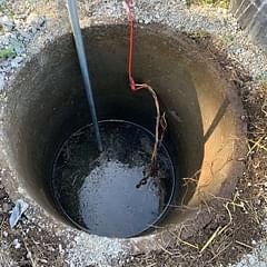 Deep well or pit with water at the bottom, red and blue pipes extending into it, surrounded by concrete and gravel