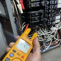 Technician holding a yellow digital multimeter in front of an electrical panel with circuit breakers and wiring